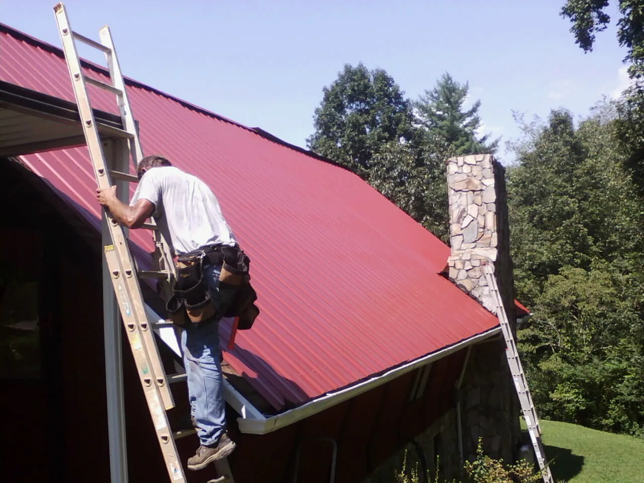Roofer on a ladder beside a red metal roof and stone chimney — Soffit & Fascia Repair by Roofing Brothers in Hagarville